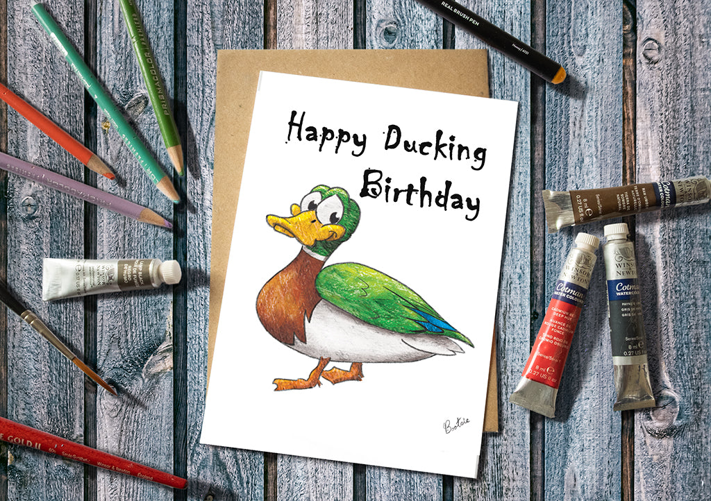Birthday card with cartoon duck and 'Happy Ducking Birthday' text, surrounded by art supplies on a wooden surface.