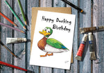 Birthday card with cartoon duck and 'Happy Ducking Birthday' text, surrounded by art supplies on a wooden surface.