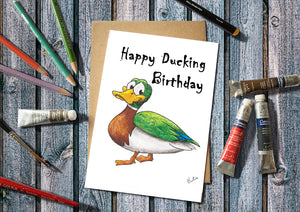 Birthday card with cartoon duck and 'Happy Ducking Birthday' text, surrounded by art supplies on a wooden surface.